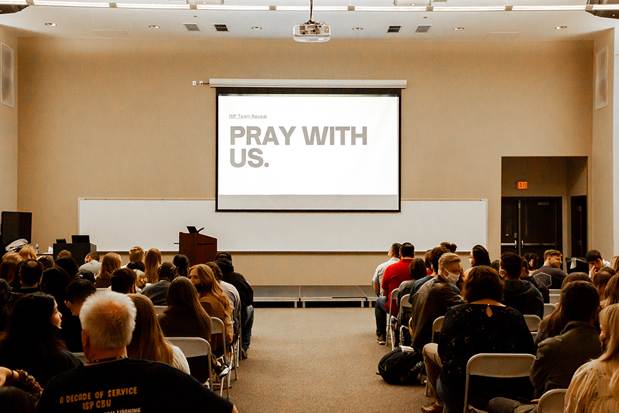 people seated in a classroom looking at a projection screen that says "PRAY FOR US"