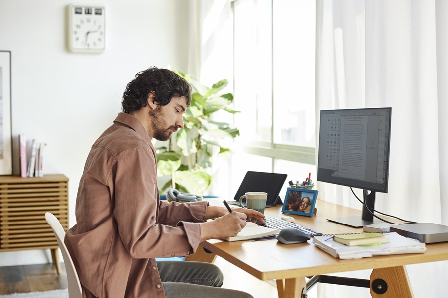 Man writing at a desk that has a computer and phone