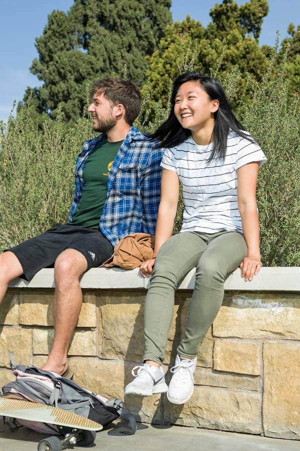 Two CBU students sitting on a stone wall by the side of a walkway on campus